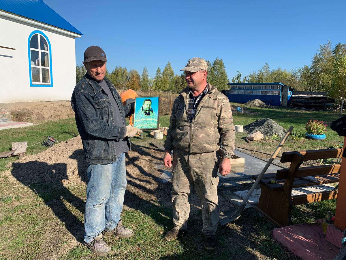 Пенсионер Александр Артеменко (держит портрет погибшего священника Максима Козачины) и каменотёс из Житомирской области Сергей ставят памятник у храма. Фото: Ольга Мусафирова, «Новая газета Европа»